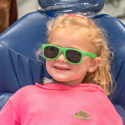 Little girl wearing sunglasses while sitting in the dentist's chair, representing representing national children’s dental health month