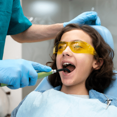 Little girl wearing yellow safety glasses while sitting in a dental chair getting teeth work done, representing pediatric dentists for special needs kids